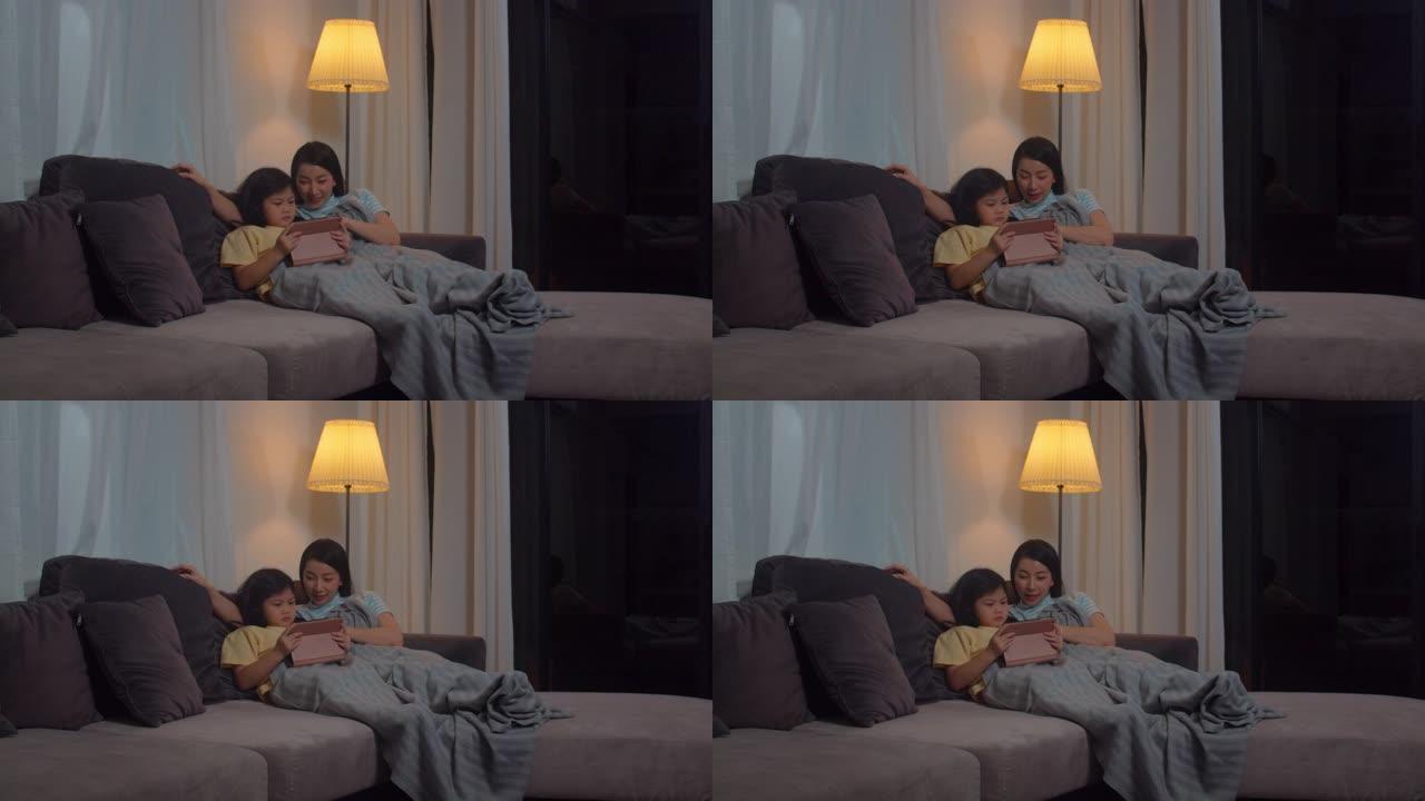 年轻的亚洲家庭和女儿在家里使用平板电脑很开心。韩国妈妈放松，小女孩躺在沙发上看电影，晚上睡在家里的客