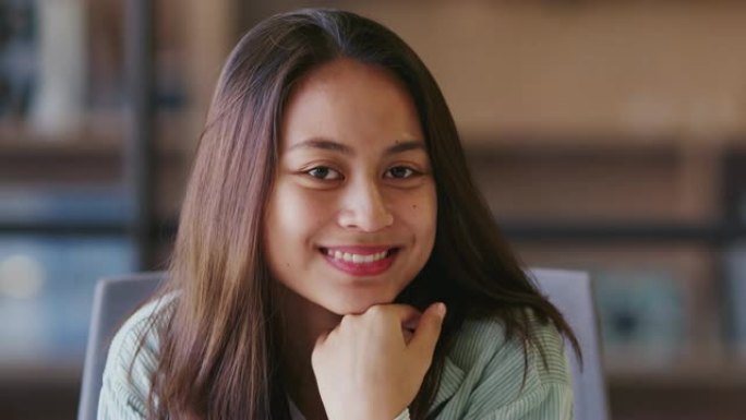 千禧一代亚洲女性创意坐在办公室里，手握下巴，对着镜头微笑，特写镜头