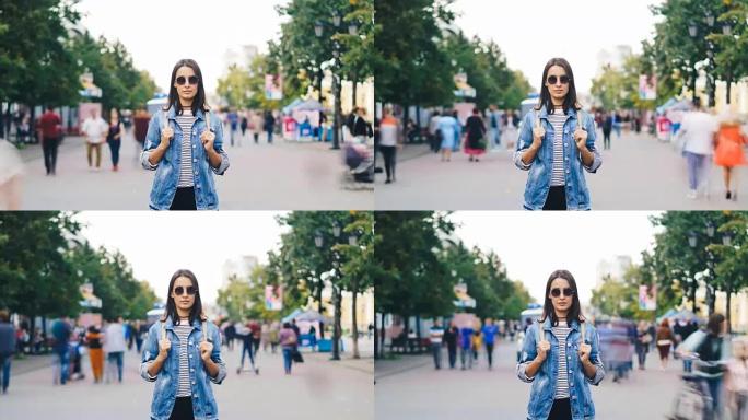 戴着墨镜的迷人黑发女人的延时肖像独自站在街上，看着秋天的男人和女人路过时的相机。
