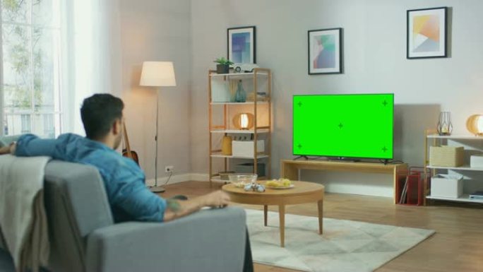在客厅里: 一个人在沙发上放松，看绿色色度键屏幕电视。