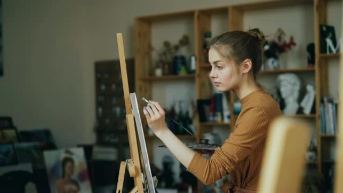 美丽的金发画家正在艺术工作室单独使用彩绘和调色板和油漆绘画。年轻女子专注于创造性的工作。
