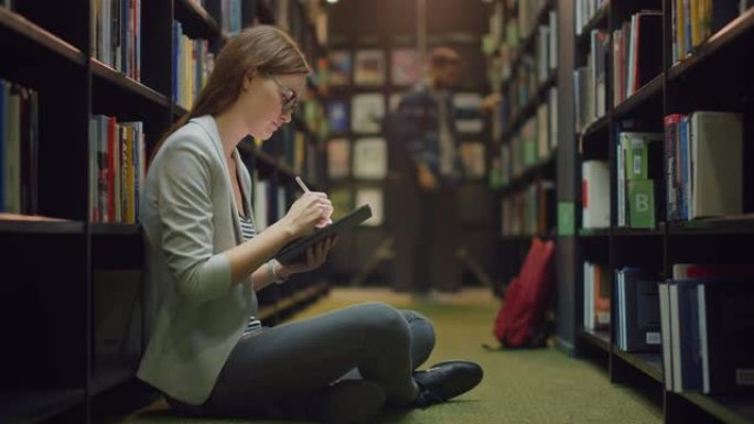 大学图书馆: 美丽的高加索女孩坐在地板上，使用数字平板电脑，写笔记，学习课堂作业。多样化的学生群体学