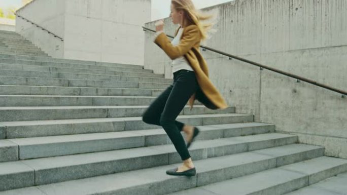 开朗快乐的年轻女子在走上楼梯时积极跳舞。她穿着一件棕色长外套。在商务中心旁边的城市混凝土公园拍摄的场