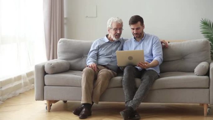 快乐的资深父亲用笔记本电脑拥抱儿子坐在沙发上