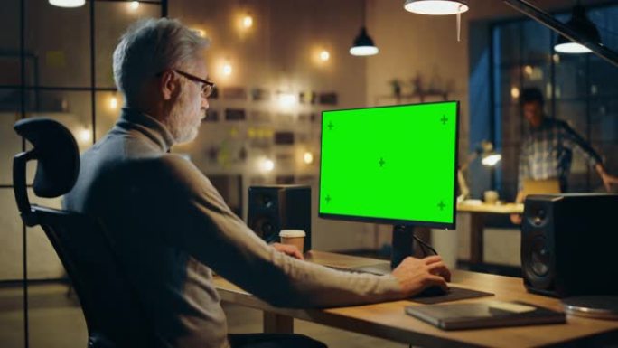 坐在办公桌前的创意中年设计师使用带有两个绿色模拟屏幕的台式计算机。专业办公室员工晚上在他的工作室工作