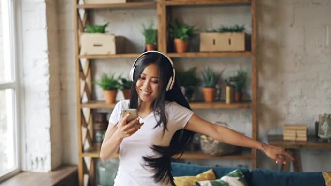 快乐的亚洲女学生的慢动作戴着耳机，拿着智能手机听音乐，跳舞，享受休闲时光。人和文化概念。