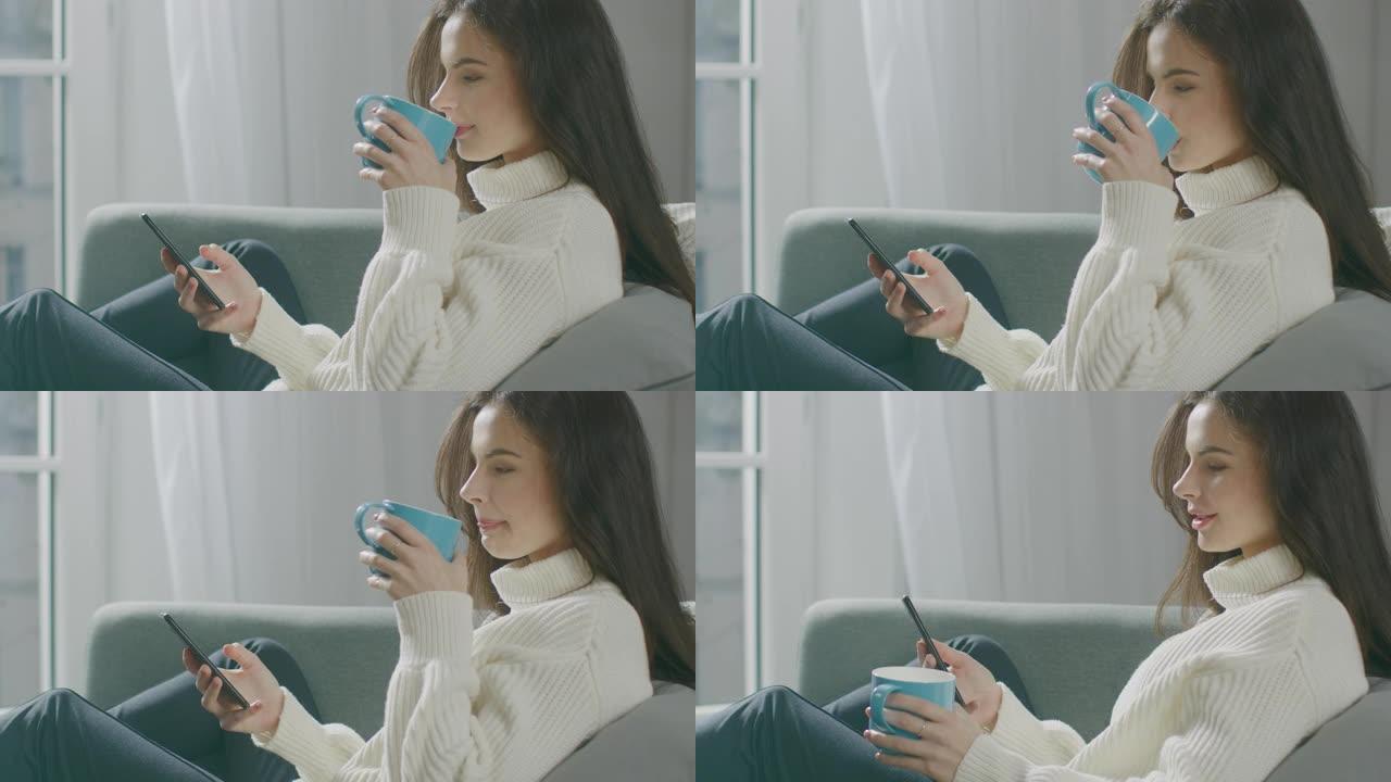 美丽的年轻女子使用智能手机，坐在椅子上喝茶。性感的女孩穿着毛衣上网，在社交媒体上发帖，在舒适的公寓里