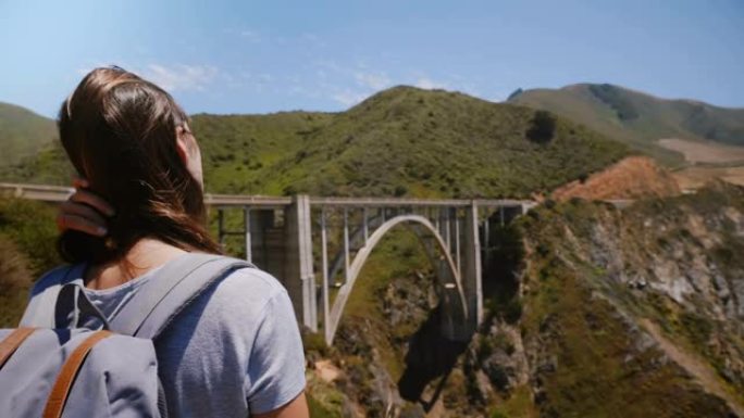 在美国大苏尔标志性的比克斯比峡谷大桥上，年轻兴奋的旅游女人背着背包观看史诗般的风景。