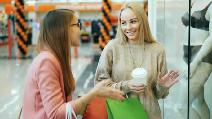 快乐的年轻女士朋友们在购物中心里看着内衣，笑着拿着外卖咖啡和纸袋，然后离开。商店和青年概念。