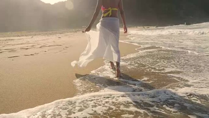 一个美丽的年轻女孩在海边漫步，穿着一顶草帽的泳衣，一条白色的方巾，金色的沙子，山峦和大海的背景。