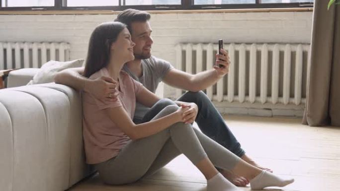 快乐的年轻夫妇拿着手机自拍坐在地板上
