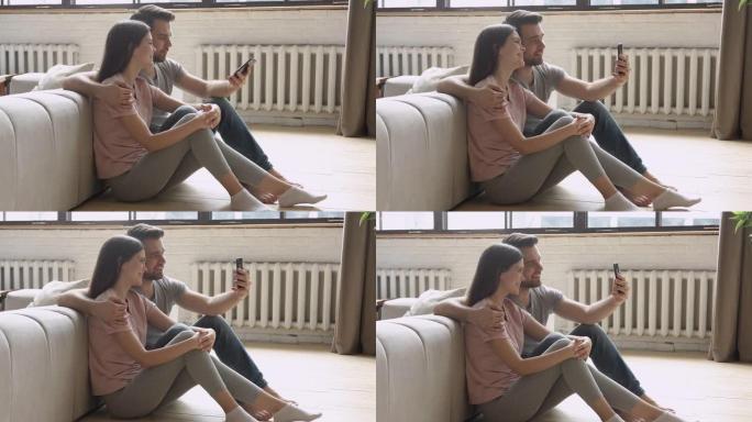 快乐的年轻夫妇拿着手机自拍坐在地板上