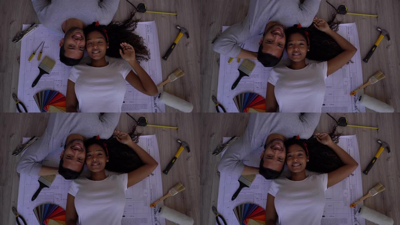 夫妇躺在蓝图上，周围有工具面对相机，在进行家庭装修后微笑