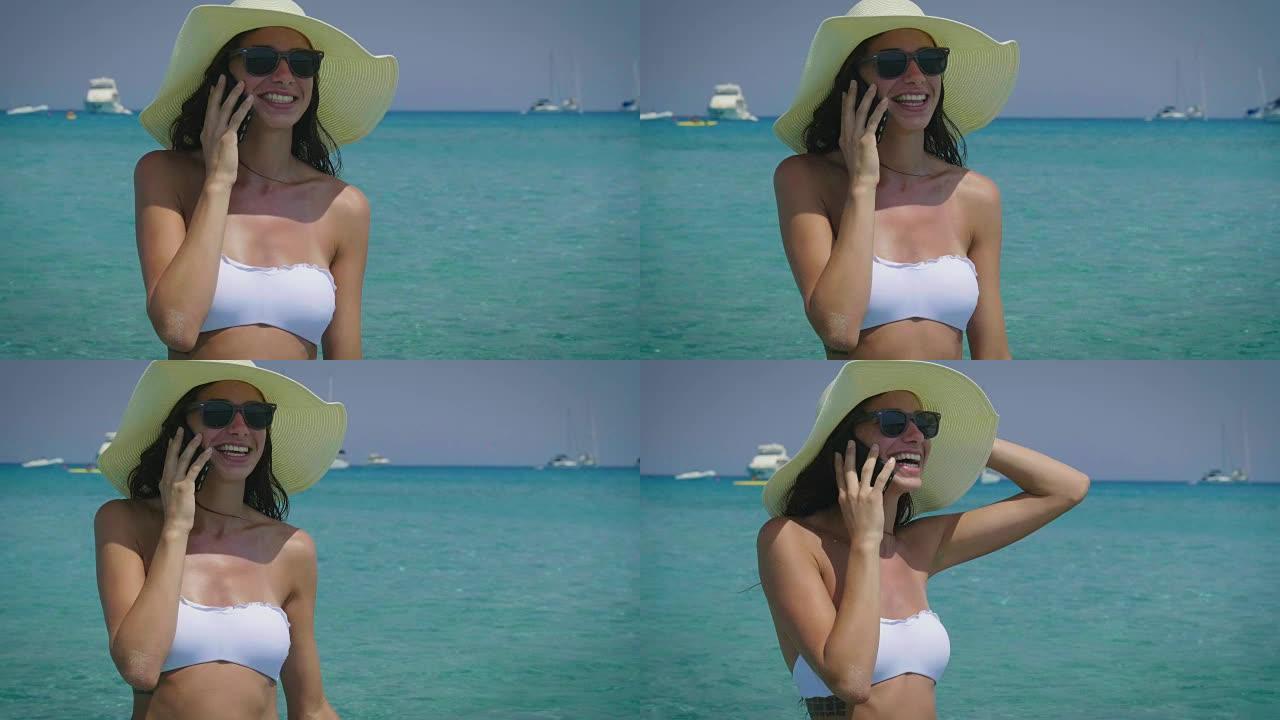 一个美丽的年轻女子微笑着，在电话里写了一条信息，白色比基尼，太阳镜，帽子，大海的背景。