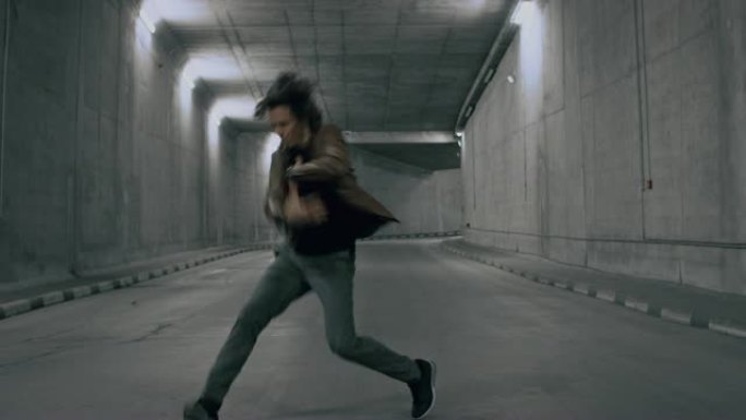 一头长发的严肃酷的年轻时髦男子正在点燃的混凝土隧道中大力跳舞嘻哈。他穿着一件棕色皮夹克。