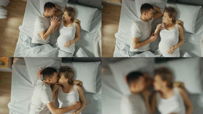 幸福的年轻夫妇在床上拥抱在一起，年轻的女人怀孕了，有爱心的丈夫温柔地抚摸着她的肚子，他们亲吻。自上而
