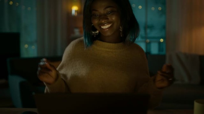 晚上: 快乐美丽的女孩坐在办公桌前跳舞，她用笔记本电脑听音乐。女孩在家玩得很开心。