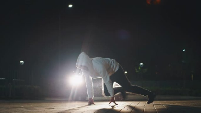 年轻的霹雳舞的慢动作很有趣，晚上在城市停车场用车灯跳舞