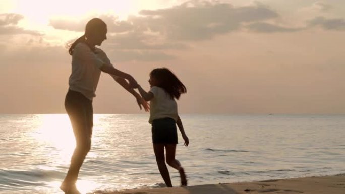 剪影快乐的母亲和女儿，在沙滩上沿着日落的边缘冲浪。活跃的父母和人们在暑假与孩子一起户外活动。家庭假期