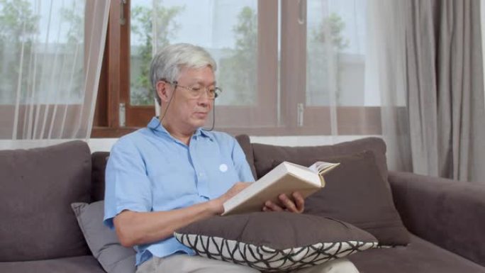 亚洲老人在家放松。亚洲高级中国男性享受休息时间看书，躺在沙发上在家概念。