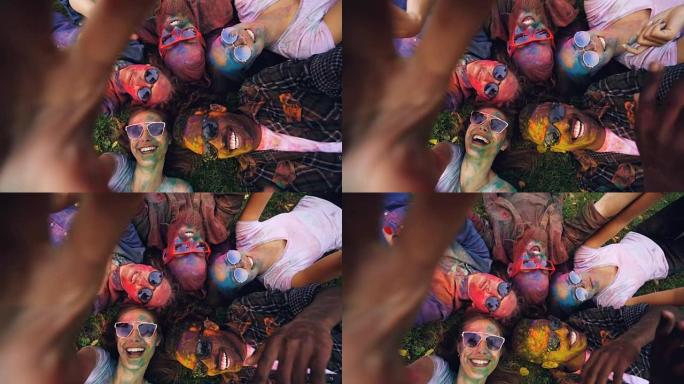 在派对胡里节上，幸福的女人和朋友躺在草地上，用相机录制自拍装置的视频。有趣和友谊的概念。
