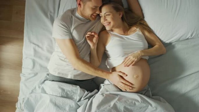 幸福的年轻夫妇在床上拥抱在一起，年轻的女人怀孕了，充满爱心的丈夫温柔地抚摸着她的肚子。顶视图摄像机。