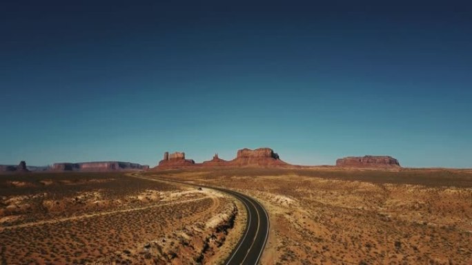 无人机在空旷的沙漠道路上升起，揭示了美国阳光明媚的纪念碑谷令人惊叹的无尽广阔空间。