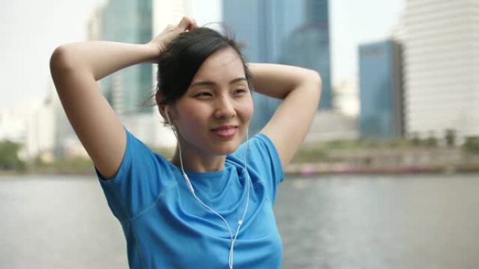年轻的亚洲健身女子在早上在城市公园跑步前拉直头发，背景是大型办公楼