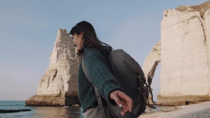 兴奋的美丽黑发旅游女人，背包旋转，双手在埃特雷塔海崖慢动作。