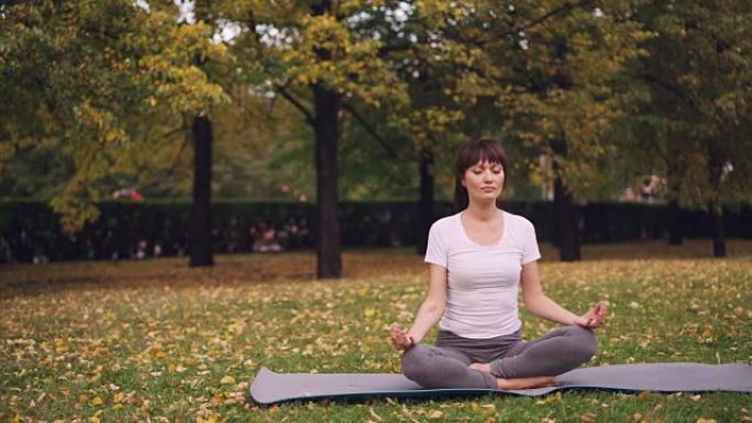 迷人的年轻女士坐在公园瑜伽垫上的莲花姿势，双手手印在膝盖上，练习后呼吸放松。冥想和自然概念。