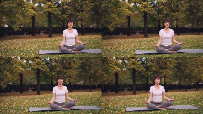 迷人的年轻女士坐在公园瑜伽垫上的莲花姿势，双手手印在膝盖上，练习后呼吸放松。冥想和自然概念。