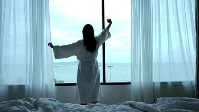 4k镜头场景迷人的亚洲女性的背面早晨醒来，在豪华酒店的卧室里锻炼并看着床上的窗户，生活方式和休闲概念