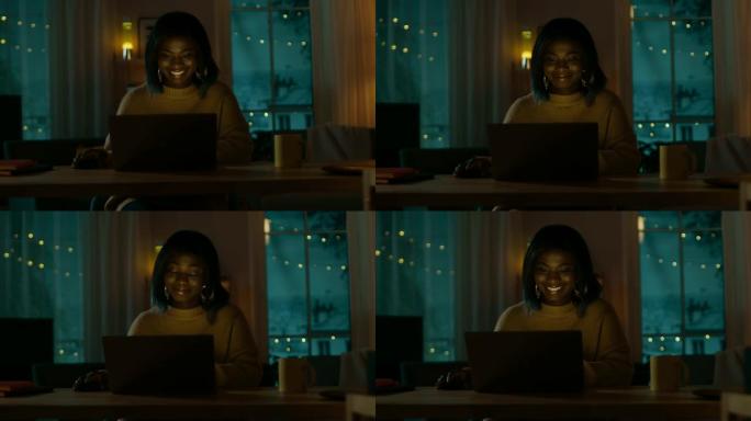 美丽微笑的黑人女孩坐在家里的办公桌前在笔记本电脑上工作的肖像。晚上，有创意的女人在舒适的客厅里用电脑