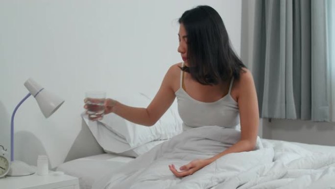 不健康患病的印度女性患有失眠。亚洲年轻女子服用止痛药缓解头痛疼痛，早上坐在家里卧室的床上喝一杯水。