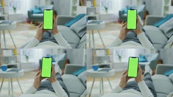 年轻女子在家使用绿色模拟屏幕智能手机。她坐在他舒适客厅的沙发上。视点摄像机拍摄。