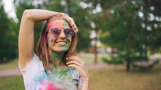 在洒红节上，迷人的年轻女子的肖像被五颜六色的油漆覆盖着微笑，看着相机，抚摸着她站在户外公园里的头发。