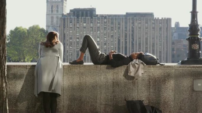 黑人千禧一代妇女躺在伦敦市泰晤士河边的堤岸墙上，与她的朋友背光交谈