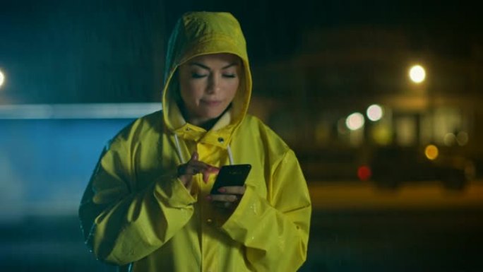 穿着黄色雨衣的美丽年轻女孩正在雨中行走和使用智能手机。她有一个温柔的微笑。天气多雨，现在是晚上。