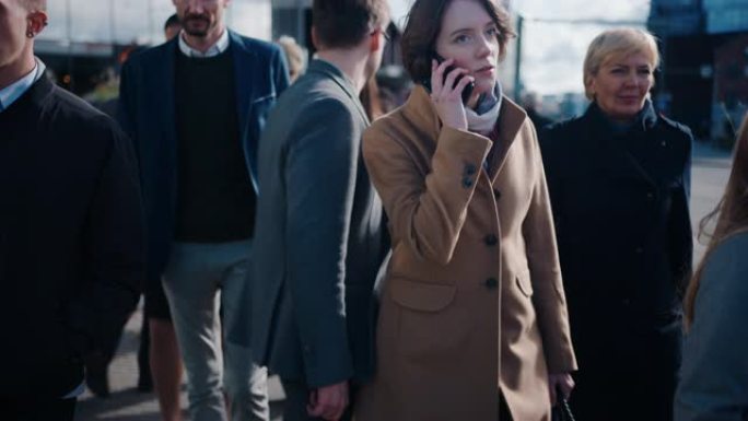 穿着棕色外套的漂亮女商人正在市中心的一条街道上用智能手机聊天。她走在拥挤的步行街上，看起来很成功。她