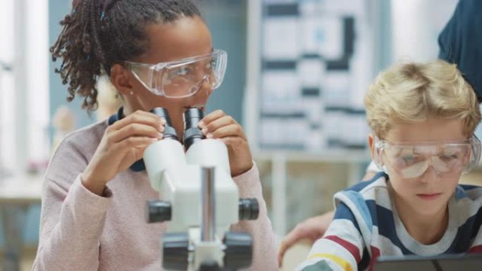 小学科学教室: 可爱的小女孩在显微镜下看，男孩在互联网上使用数字平板电脑检查信息。老师从后面观察