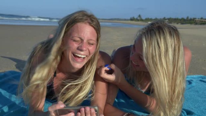 南非海滩上，两个美丽的年轻女子躺在毛巾上，在手机上笑着分享笑话的4k前视图