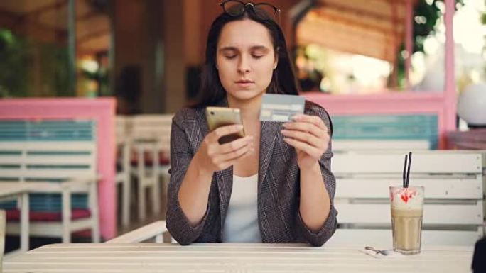 漂亮的年轻女子拿着银行卡和智能手机进行在线支付触摸屏然后微笑。网络购物和现代技术概念。