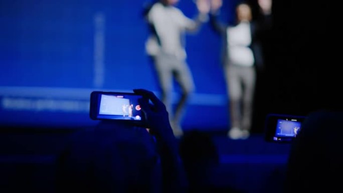 观众使用智能手机在商务会议上录制两名演讲者。在观众厅公众使用手机拍摄两位明星主持人的视频，现场活动。