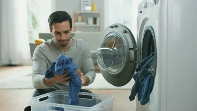 穿着灰色牛仔裤和外套的英俊严肃的年轻人坐在家里的洗衣机前。他给洗衣机装上脏衣服。明亮宽敞的客厅，室内
