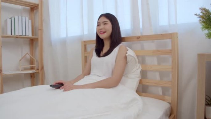 年轻的亚洲女人在家看电视，女人躺在卧室的床上感到快乐。生活方式女人早上在家放松的概念。慢动作镜头。