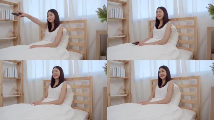 年轻的亚洲女人在家看电视，女人躺在卧室的床上感到快乐。生活方式女人早上在家放松的概念。慢动作镜头。