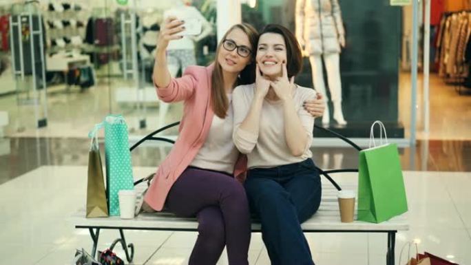漂亮的年轻女性正坐在购物中心的长凳上玩得开心，使用智能手机相机自拍。现代科技、商店和朋友概念。