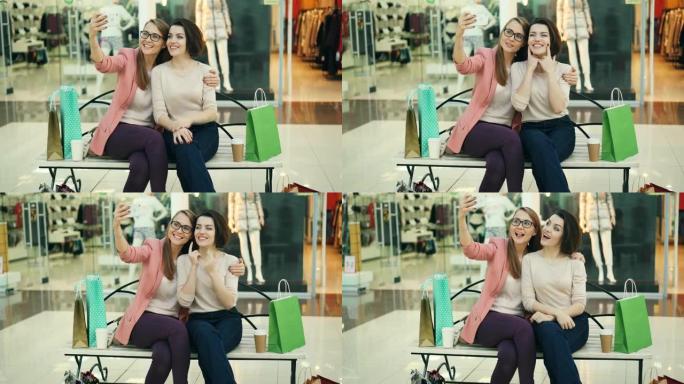 漂亮的年轻女性正坐在购物中心的长凳上玩得开心，使用智能手机相机自拍。现代科技、商店和朋友概念。