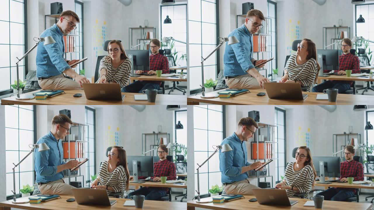 男同事坐在一个年轻的女Cowroker面前的桌子上，拿着一台带有商务笔记的平板电脑。创意机构的友好氛