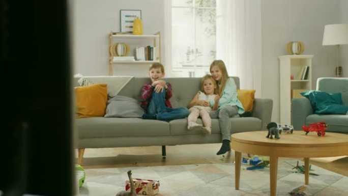 两个可爱的小女孩和年轻可爱的男孩坐在沙发上看电视。快乐的孩子在电视上看卡通片，在客厅里笑。